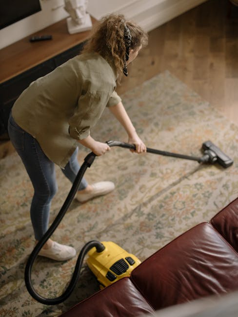 A young boy using a vacuum cleaner for surface cleaning on an orange and beige rug in a well-lit living room with large windows, cream-colored sofas with cushions, and polished wooden flooring. An old