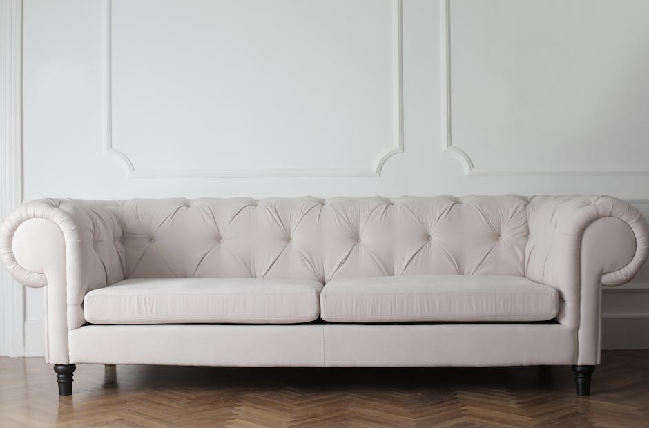 A white tufted fabric sofa with rolled arms and black wooden legs is positioned against a white wall with decorative moldings. The sofa appears clean, well-maintained, and free of stains, situated in 